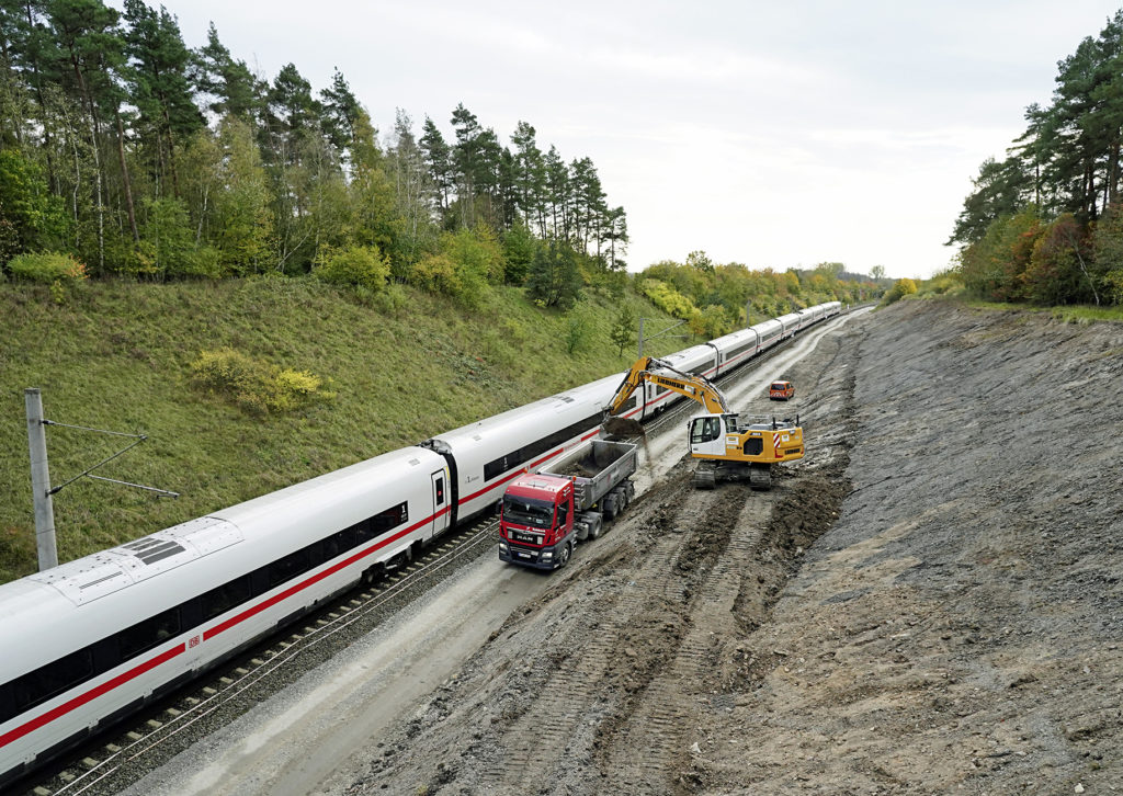 ICE fährt neben einer Gleisbaustelle vorbei, LKW und Bagger stehen daneben