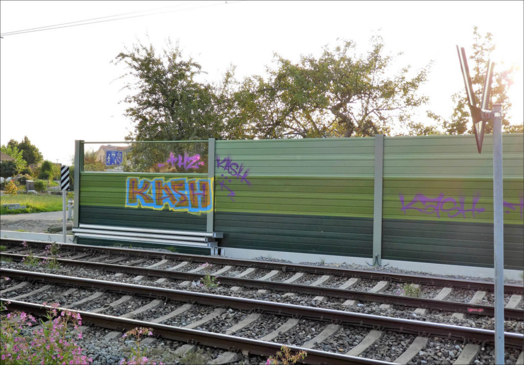 Grüne Schallschutzwand mit Graffiti verschmiert, im Vordergrund zwei Gleise