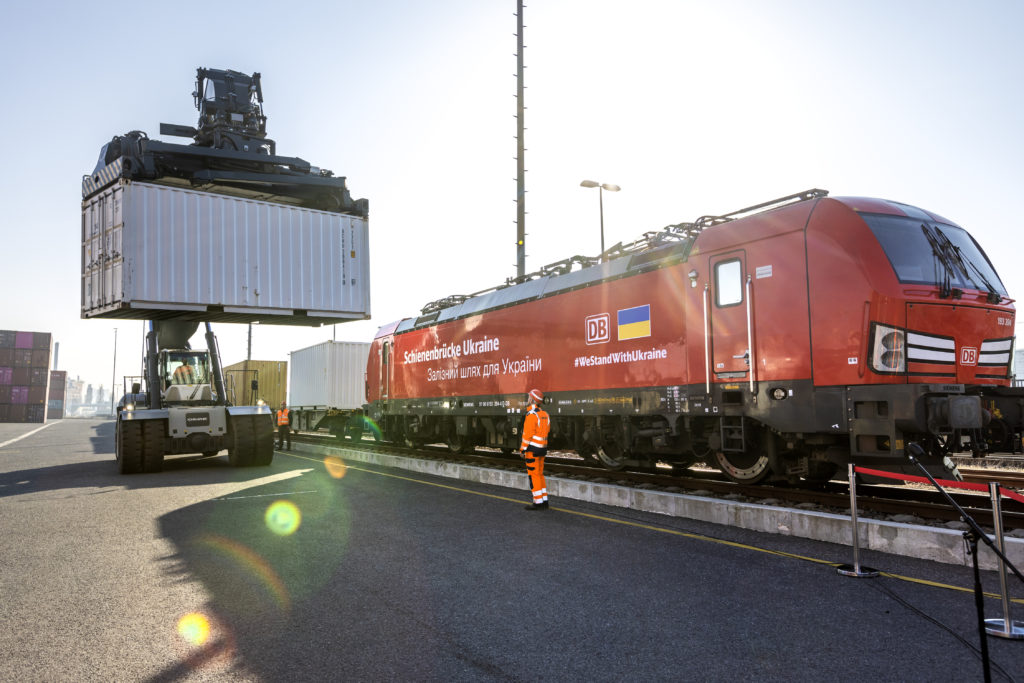 Kran mit Container vor einem Zug mit der Aufschrift Schienenbrücke Ukraine