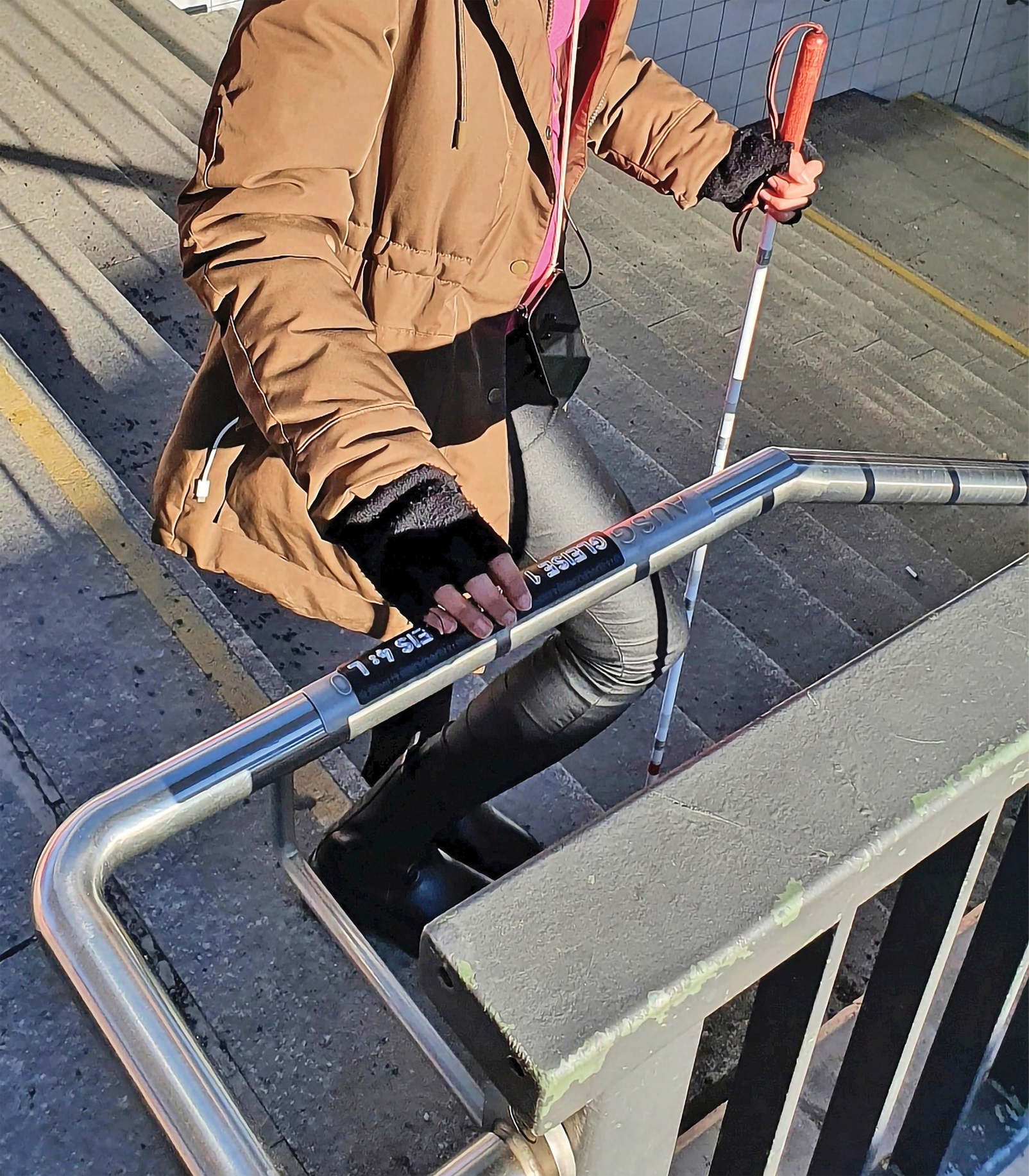 Eine blinde Frau ertastet im Bahnhof München-Pasing das Handlaufschild am Geländer einer Treppe. Zum Vergleich ist ein kontrastreiches Schild einfach auf das bisherige Schild aufgelegt worden. Sowohl am Beginn als auch am Ende des Treppenhandlaufs sind an jedem Geländer die Informationen angebracht.